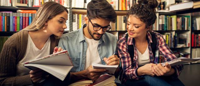 Diverse group of students in library conversing with pencils, literature and books in hand