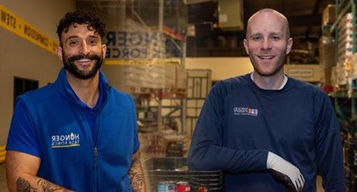 Two smiling volunteers in blue shirts, one reading "feed more" and the other "hunger task force," standing in a food bank warehouse.